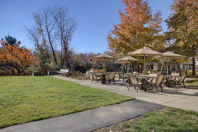 Outdoor patio area with several tables and chairs under beige umbrellas, surrounded by green grass and trees with autumn foliage. A paved walkway runs alongside the patio, and a white bench is visible near the trees.
