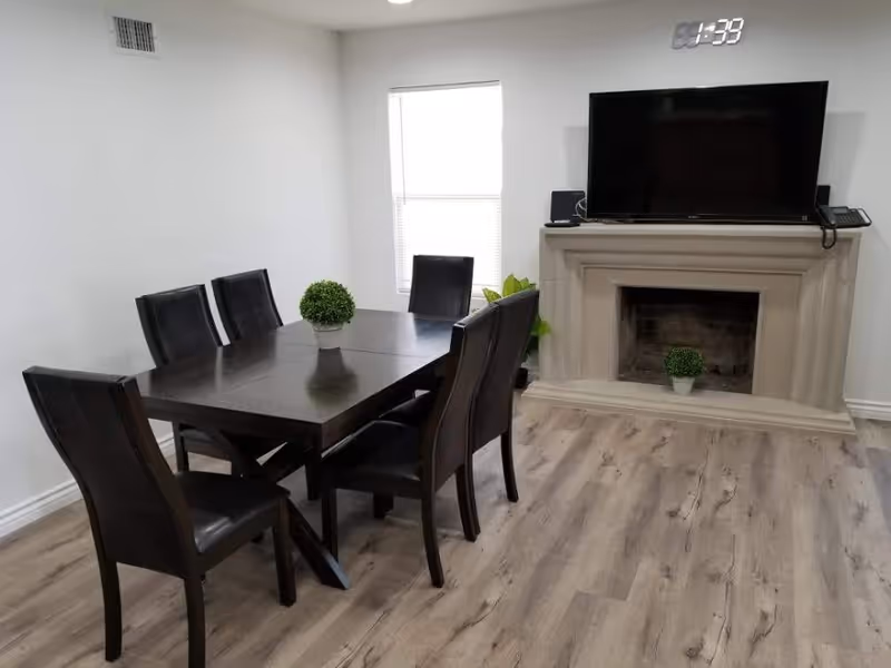 A dining room with a dark wooden table surrounded by six black leather chairs. A small green plant is placed in the center of the table. There is a window with blinds on the back wall, and a fireplace with a TV mounted above it. Another small green plant is placed inside the fireplace. The room has light-colored wooden flooring and white walls.