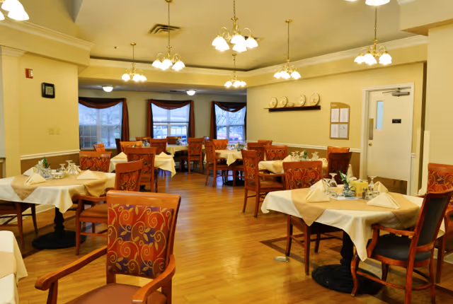 A dining room with multiple tables covered with white and beige tablecloths, each set with napkins and small centerpieces. The room has wooden floors, several wooden chairs with red patterned upholstery, and large windows with red valances. Ceiling lights hang from the ceiling, illuminating the space warmly.