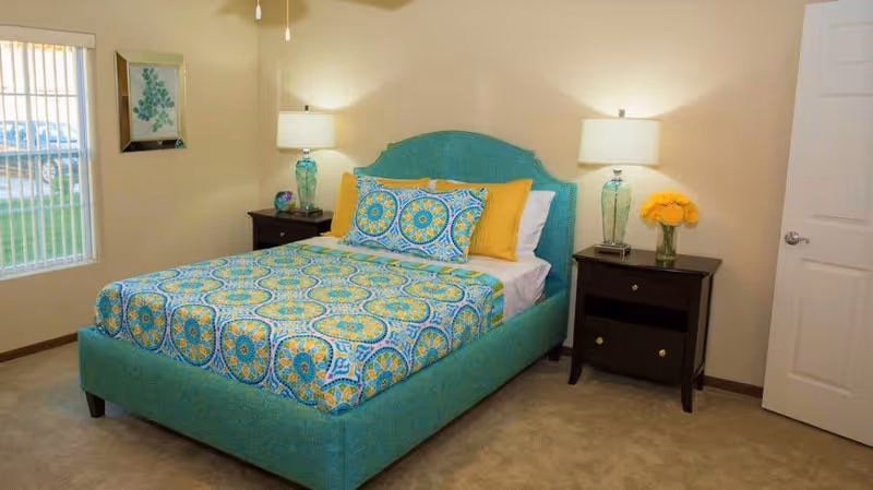 A bedroom with a turquoise upholstered bed featuring a colorful patterned bedspread and pillows in blue, yellow, and white. There are two dark wooden nightstands on either side of the bed, each with a lamp and decorative items. A window with vertical blinds is on the left wall, and a closed white door is on the right.