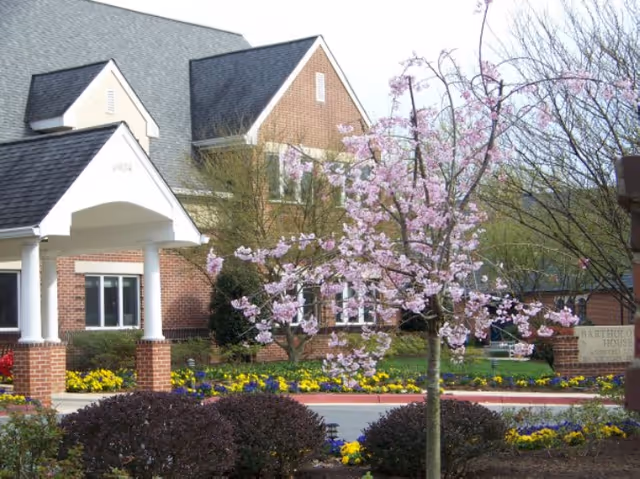 Brick building entrance with a white portico, a blooming pink tree, and colorful landscaped flower beds in front.