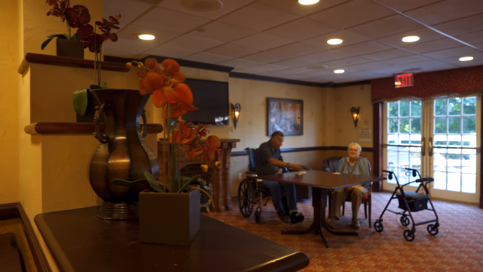 A cozy common area in a senior living facility with two elderly individuals sitting at a table. One person is in a wheelchair and the other is seated on a chair. A walker is nearby. The room has warm lighting, a TV mounted on the wall, a framed picture, and large windows with glass doors letting in natural light. In the foreground, there is a table with decorative vases and orange flowers.
