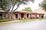 Exterior view of a single-story brick building with a covered porch area supported by columns. There are several chairs on the porch and a large tree in the front yard. The building is adjacent to a paved road with a grassy strip and a sidewalk.
