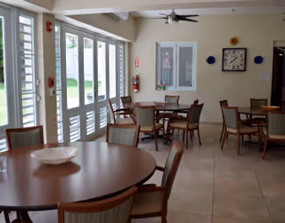 A bright dining room with several round wooden tables and chairs arranged neatly. Large windows on the left side let in natural light, and there is a fire extinguisher mounted on the wall. A clock and decorative items are hung on the far wall.