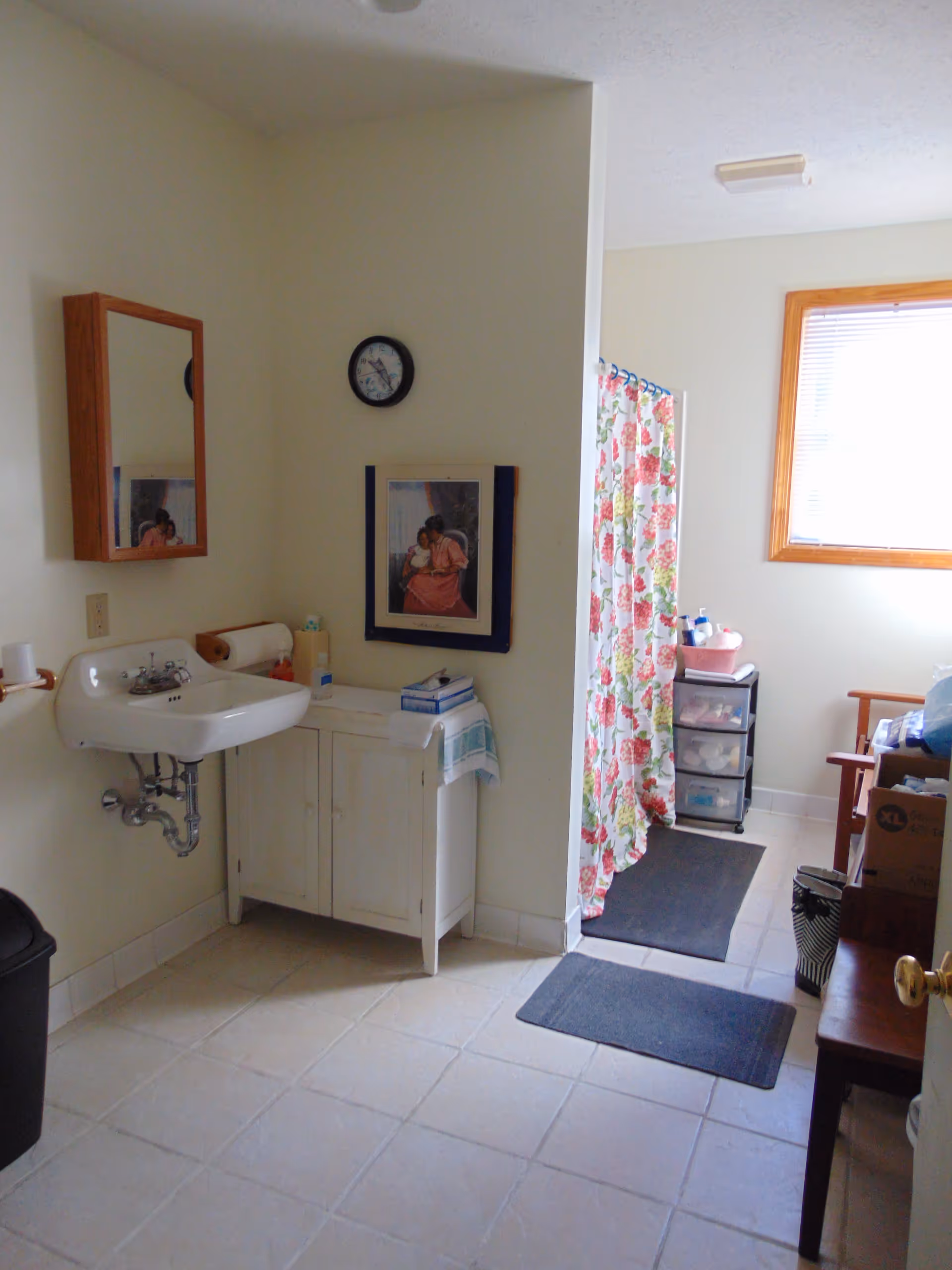 A bathroom with a wall-mounted sink, a wooden medicine cabinet with a mirror, a clock, and a framed picture on the wall. There is a white cabinet below the picture, a floral shower curtain, two black floor mats, a window with blinds, and various toiletries on a small plastic drawer unit. A wooden bench and some boxes are also visible.