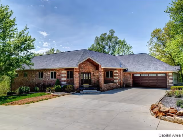 A single-story brick house with a gray shingled roof, a large driveway, and a two-car garage. The house is surrounded by green trees and shrubs under a clear blue sky.