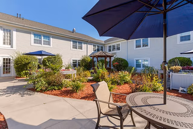 Sunlit courtyard with patio tables and umbrellas, landscaped flower beds and a small gazebo surrounded by a two-story building.