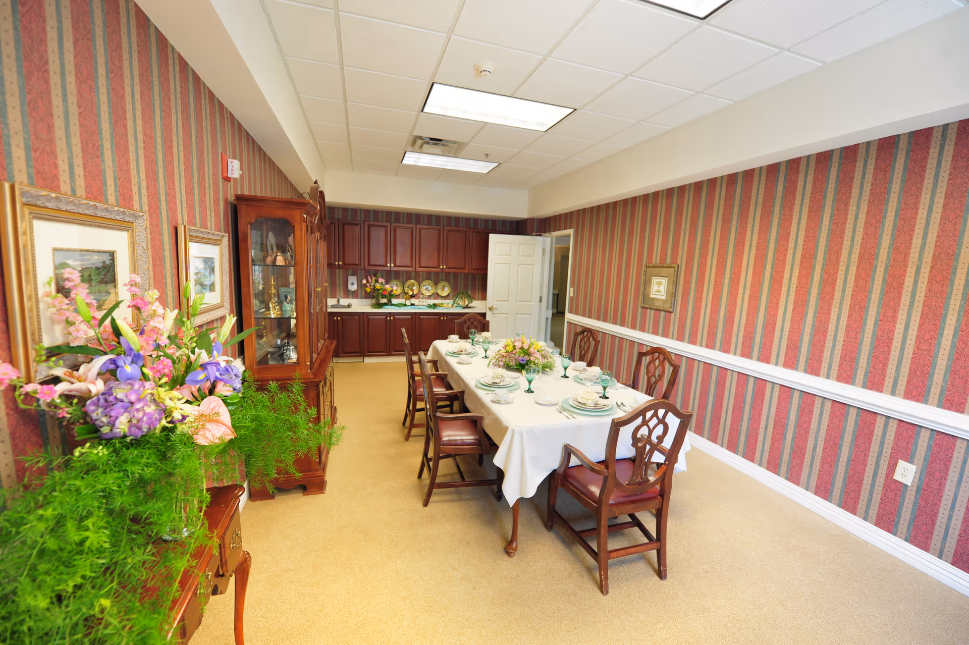 A formal dining room with a long table covered with a white tablecloth, set with plates, glasses, and floral centerpieces. The room has striped wallpaper in red and gold tones, wooden chairs with cushioned seats, a wooden china cabinet, framed artwork on the walls, and a sideboard with a large floral arrangement. The back of the room features dark wooden cabinets and a door leading to another area.