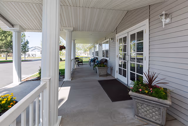 Covered porch area of a senior living facility with white columns, gray siding, potted plants, and several chairs along the walkway. The porch overlooks a green lawn and a driveway.