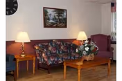 A seating area with a floral sofa, wooden coffee table topped with a flower arrangement, two table lamps, a wingback chair, wall clock, and a landscape painting.