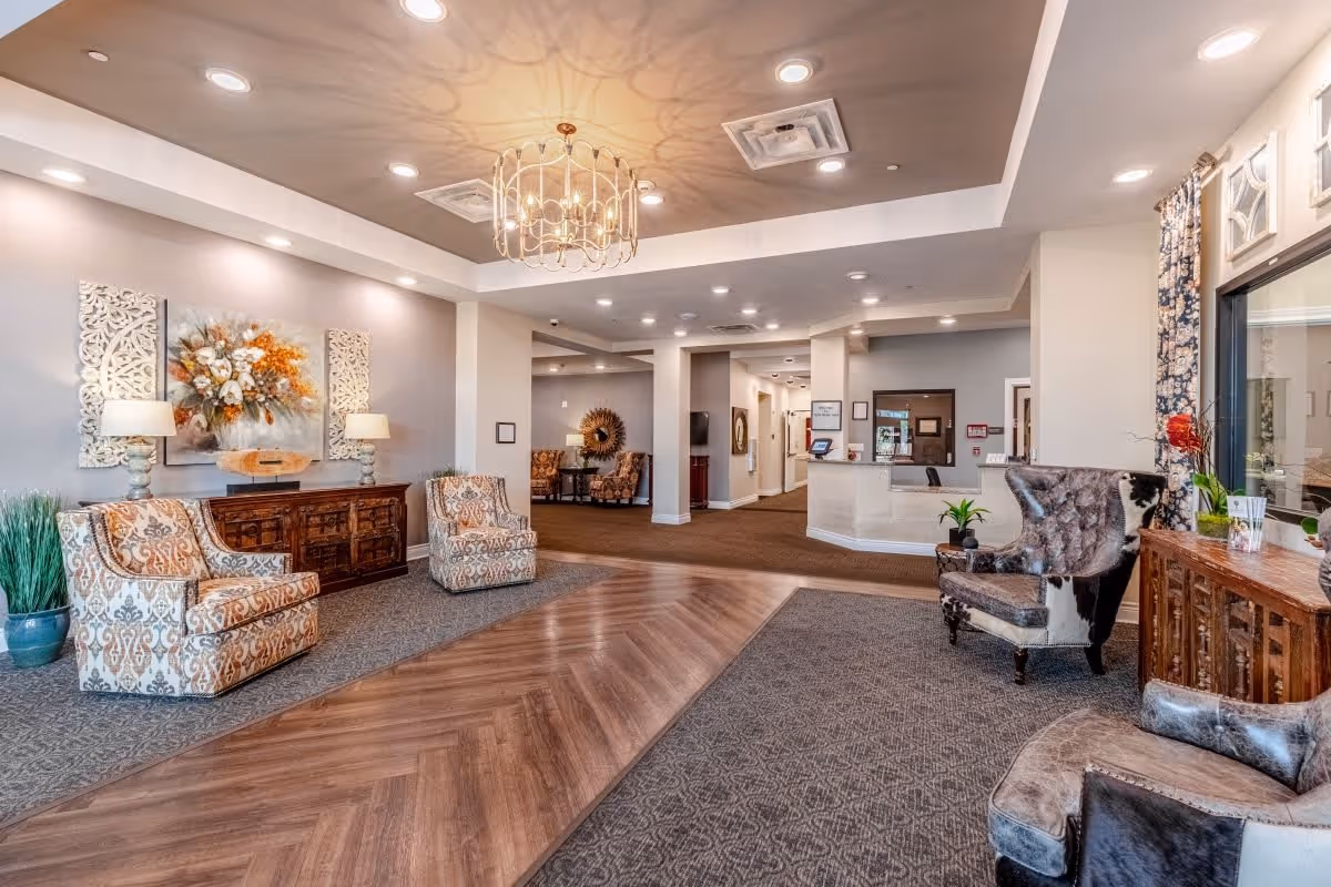 A spacious and well-lit senior living facility lobby with patterned armchairs, a wooden sideboard with lamps and floral artwork, a chandelier casting decorative shadows on the ceiling, and a reception desk in the background. The floor features a combination of wood and carpet, and there are plants and decorative curtains near the windows.