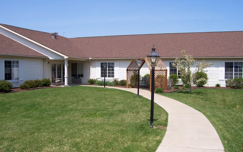 Single-story building with white siding and a brown roof, surrounded by a well-maintained lawn. A curved concrete walkway leads to the entrance, passing by two black lamp posts and a wooden lattice structure with benches underneath. Small bushes and trees are planted along the building's exterior.