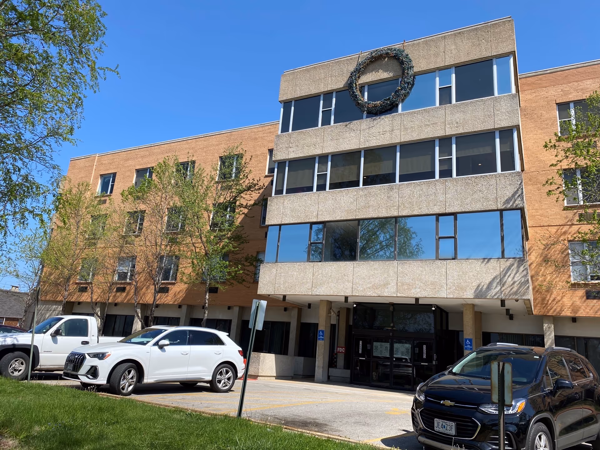 Front exterior of a four-story brick and concrete senior living building with a large wreath above the entrance and several cars parked in front.