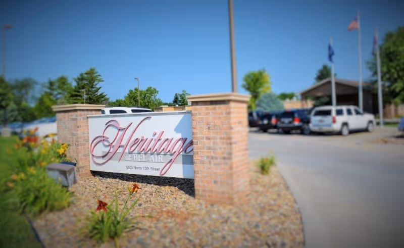 Outdoor view of the entrance sign for Heritage of Bel-Air, mounted on a brick structure with landscaping and flowers around it. Several parked cars and a building with flagpoles are visible in the background under a clear blue sky.