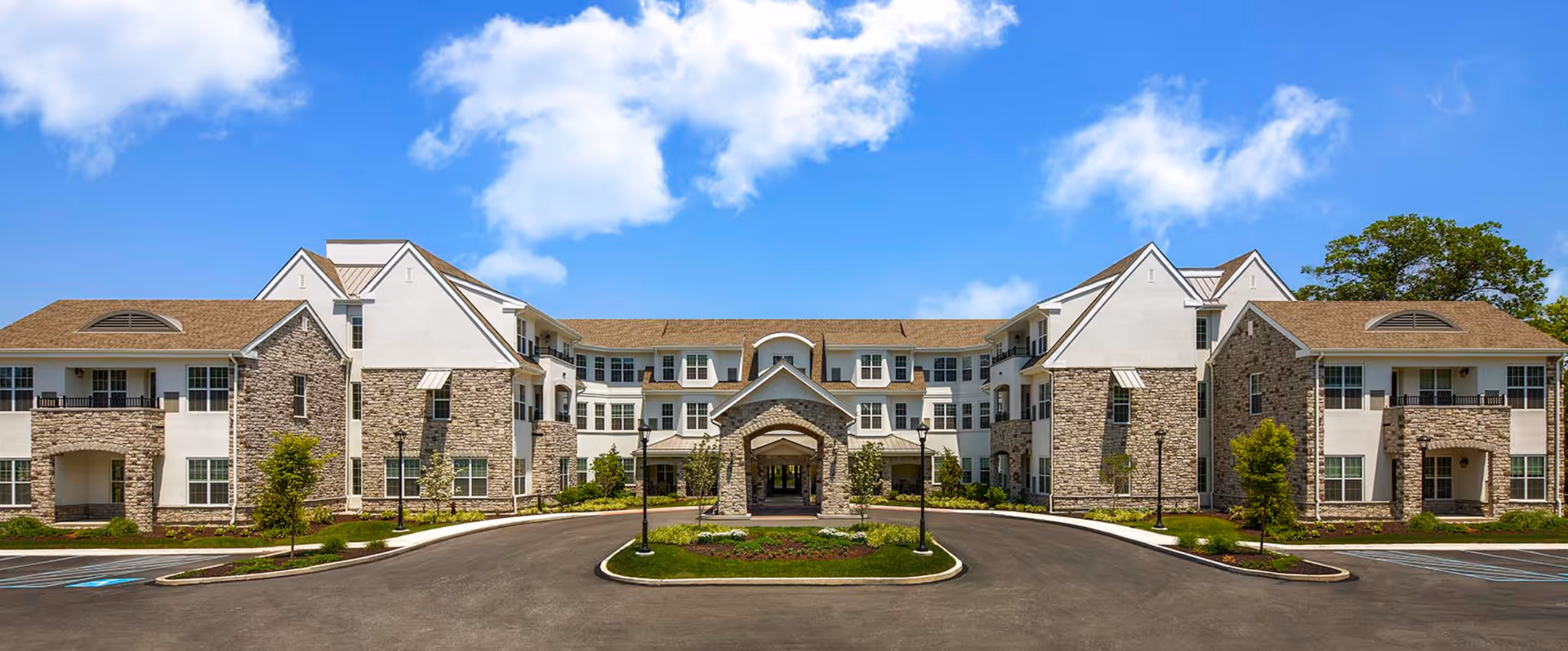 Front exterior view of Atria Lafayette Hill facility showing a large, multi-story building with stone and white siding, multiple windows, and a covered entrance. The sky is blue with some clouds, and there is a paved driveway and landscaped greenery in front.