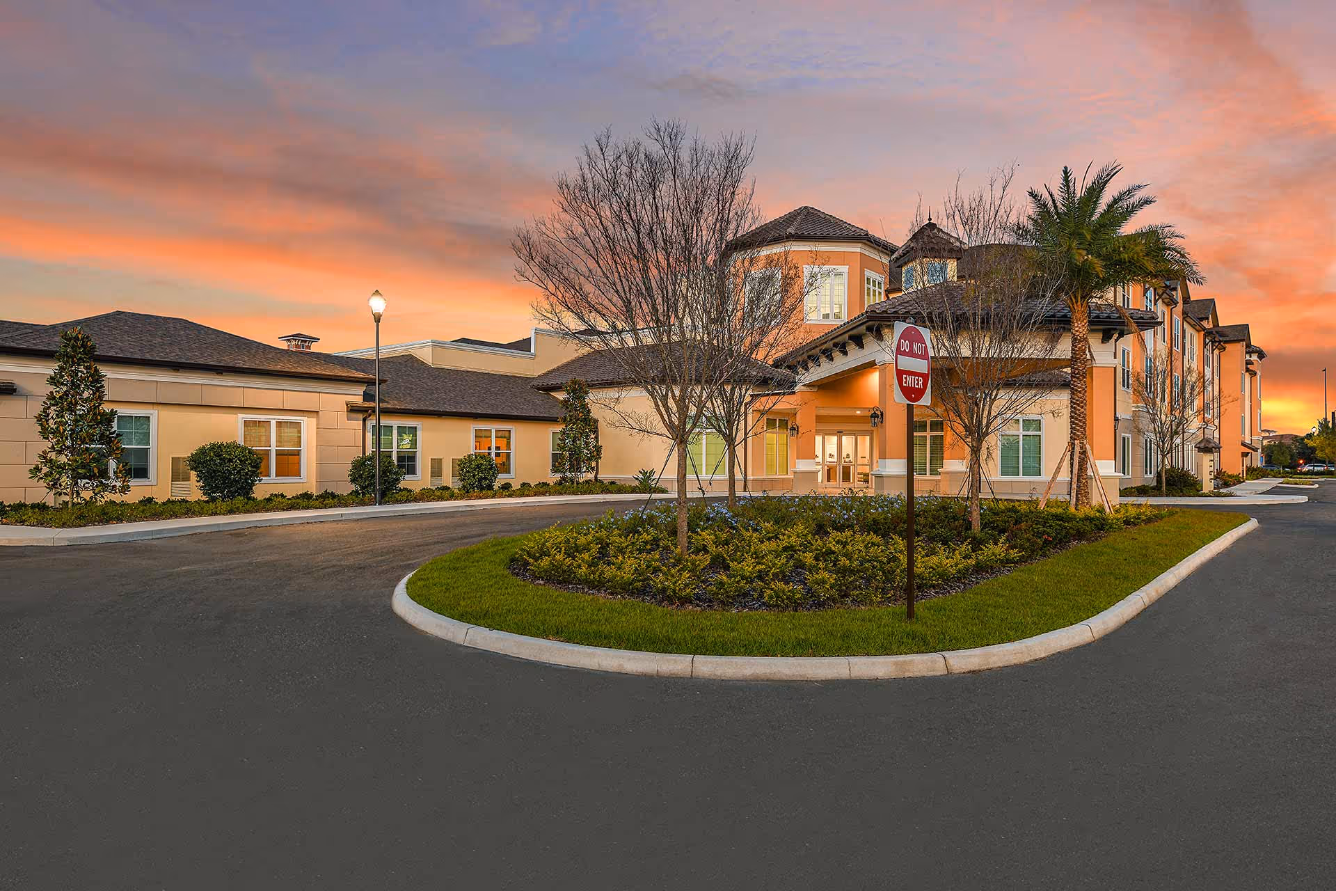 Front exterior of a senior living facility with a circular driveway, landscaped island, palm tree and a 'Do Not Enter' sign at sunset.