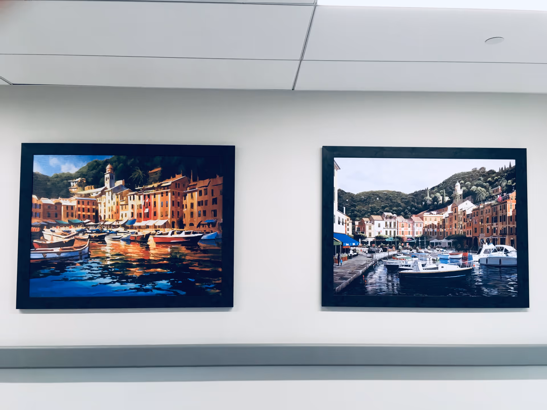 Two framed paintings hanging on a white wall above a gray handrail. Both paintings depict colorful coastal village scenes with boats docked in the water and buildings in the background.
