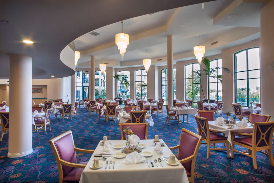 A spacious dining room with multiple tables set with white tablecloths, napkins, plates, cups, and silverware. The room features large windows allowing natural light to enter, purple cushioned chairs, decorative plants, and elegant chandeliers hanging from the ceiling.
