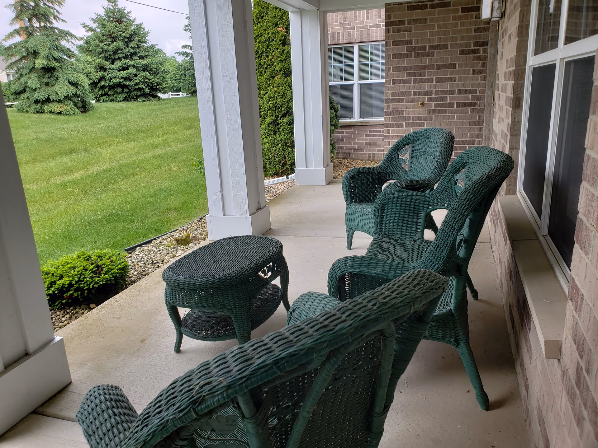 A covered outdoor patio area with green wicker chairs and a matching wicker table. The patio overlooks a grassy lawn with trees and shrubs, and the building exterior is made of brick with white pillars supporting the roof.