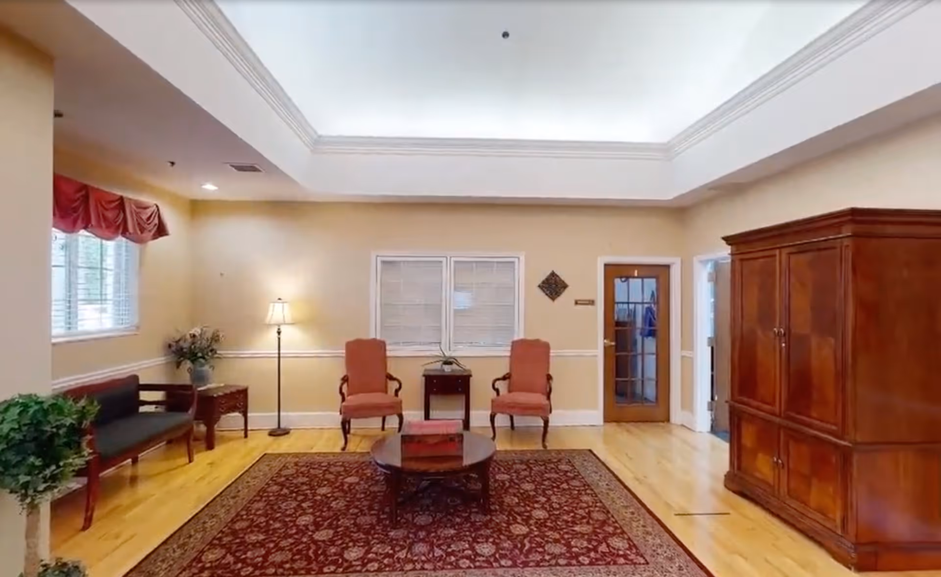 A cozy living room area with wooden flooring, a large patterned rug, two upholstered armchairs, a round wooden coffee table, a wooden bench with dark cushions, a floor lamp, a potted plant, and a large wooden cabinet. The room has beige walls, a window with blinds, and a door with glass panels.