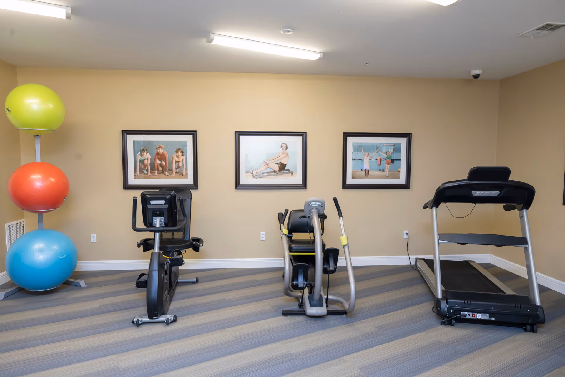 A small exercise room with three pieces of cardio equipment including a stationary bike, a recumbent bike, and a treadmill. To the left, there is a rack holding three large exercise balls in green, orange, and blue. The walls are beige and decorated with three framed pictures of people exercising. The floor has a striped pattern in shades of gray and blue.