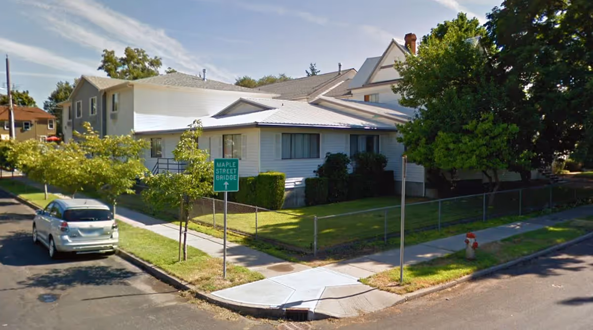 Exterior view of a white multi-section building with a sloped roof, surrounded by a green lawn, small trees, and a chain-link fence. A silver car is parked on the street next to the sidewalk, and a green street sign indicates the direction to Maple Street Bridge. The sky is partly cloudy.