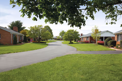 A paved road curves through a residential area with single-story brick buildings on either side, surrounded by well-maintained green lawns and trees under a partly cloudy sky.