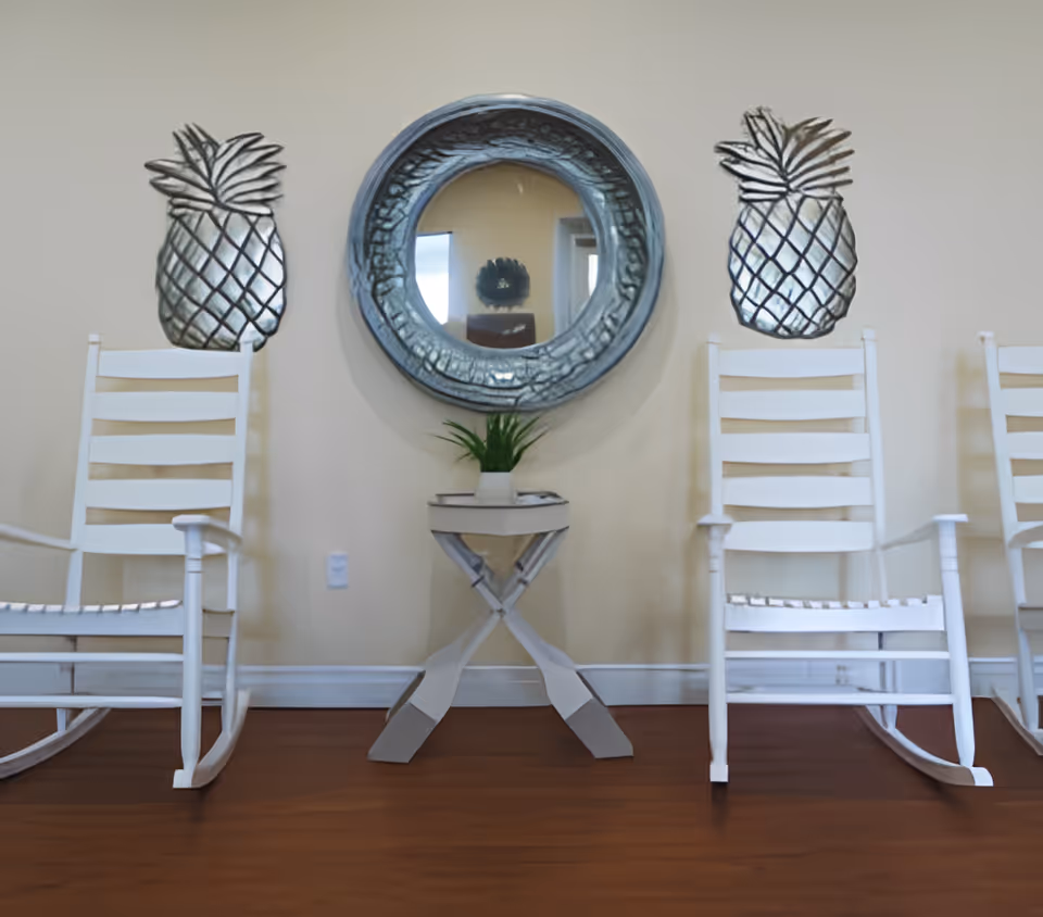 Interior view of a room with two white wooden rocking chairs on either side of a small white table with a green plant on top. A round decorative mirror hangs on the wall above the table, flanked by two metal pineapple wall decorations. The floor is wooden and the walls are light-colored.