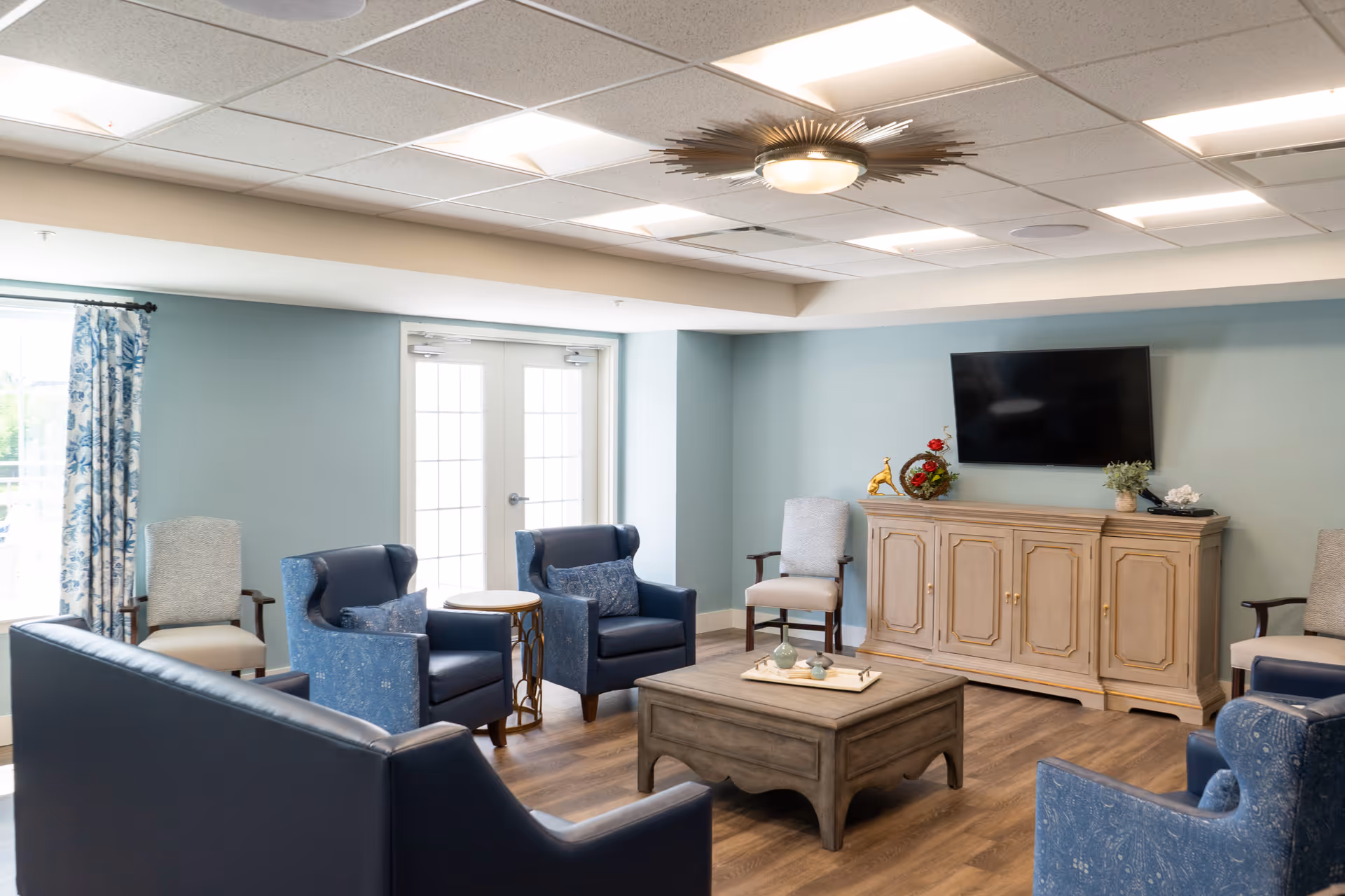 A bright and cozy living room with blue and beige armchairs arranged around a wooden coffee table. There is a wooden cabinet with decorative items and a flat-screen TV mounted on the wall above it. Large windows with floral curtains allow natural light to fill the room, and the ceiling features a modern light fixture.