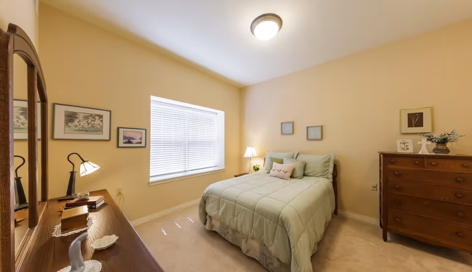 A cozy bedroom with beige walls and carpeted floor. The room features a bed with light green bedding and multiple pillows, a wooden dresser with decorative items, a wooden vanity with a mirror and a lamp, and a window with white blinds letting in natural light.