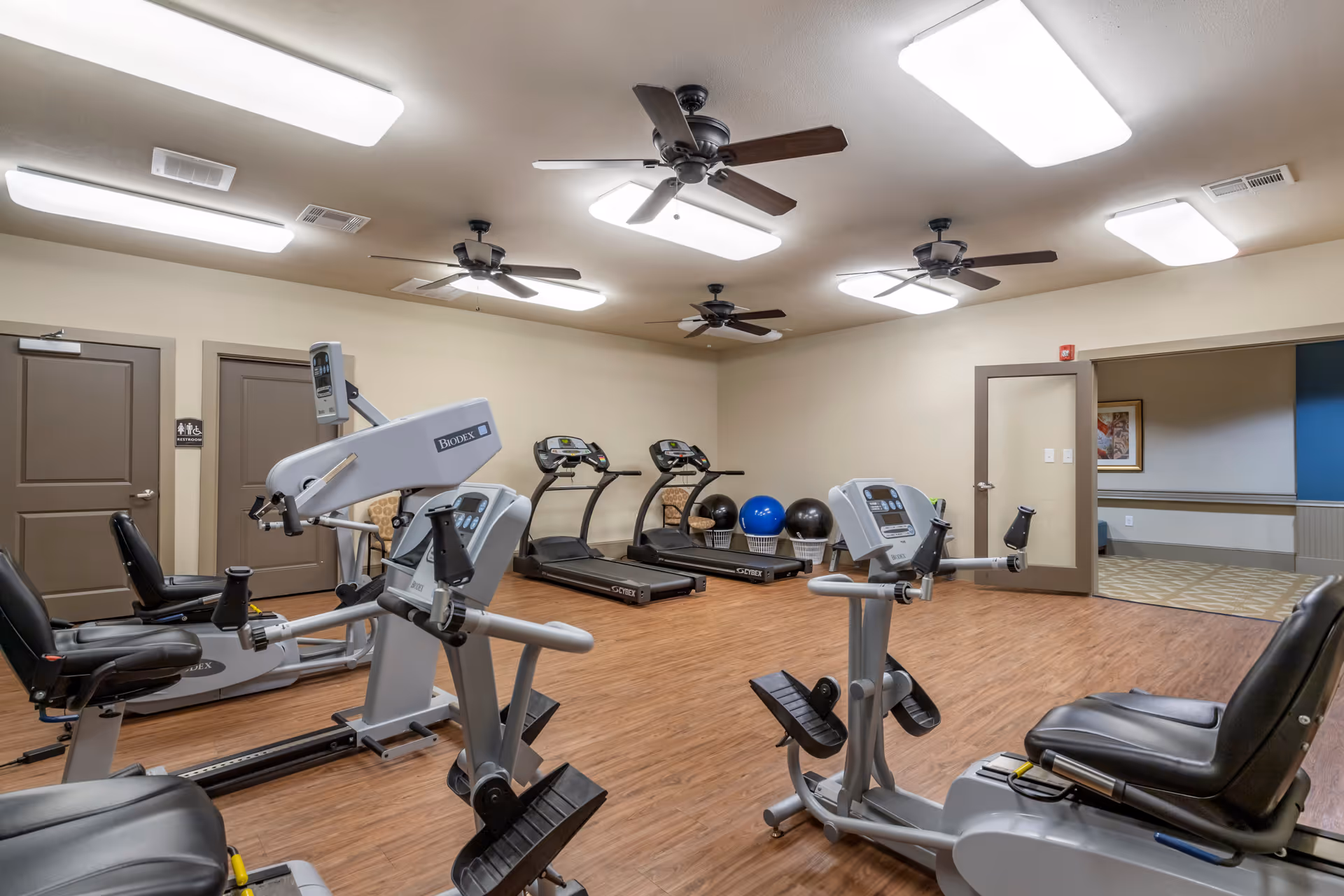 A fitness room in a senior living facility with exercise equipment including recumbent bikes and treadmills. The room has wood flooring, beige walls, ceiling fans, and bright overhead lighting. There are exercise balls and a doorway leading to another room.