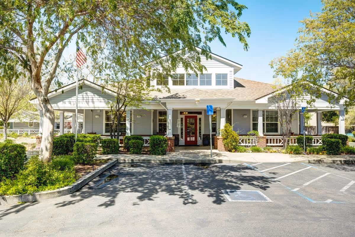 Front exterior view of a single-story building with a covered porch, surrounded by trees and bushes, with a parking lot and handicap parking spaces in front under a clear blue sky.