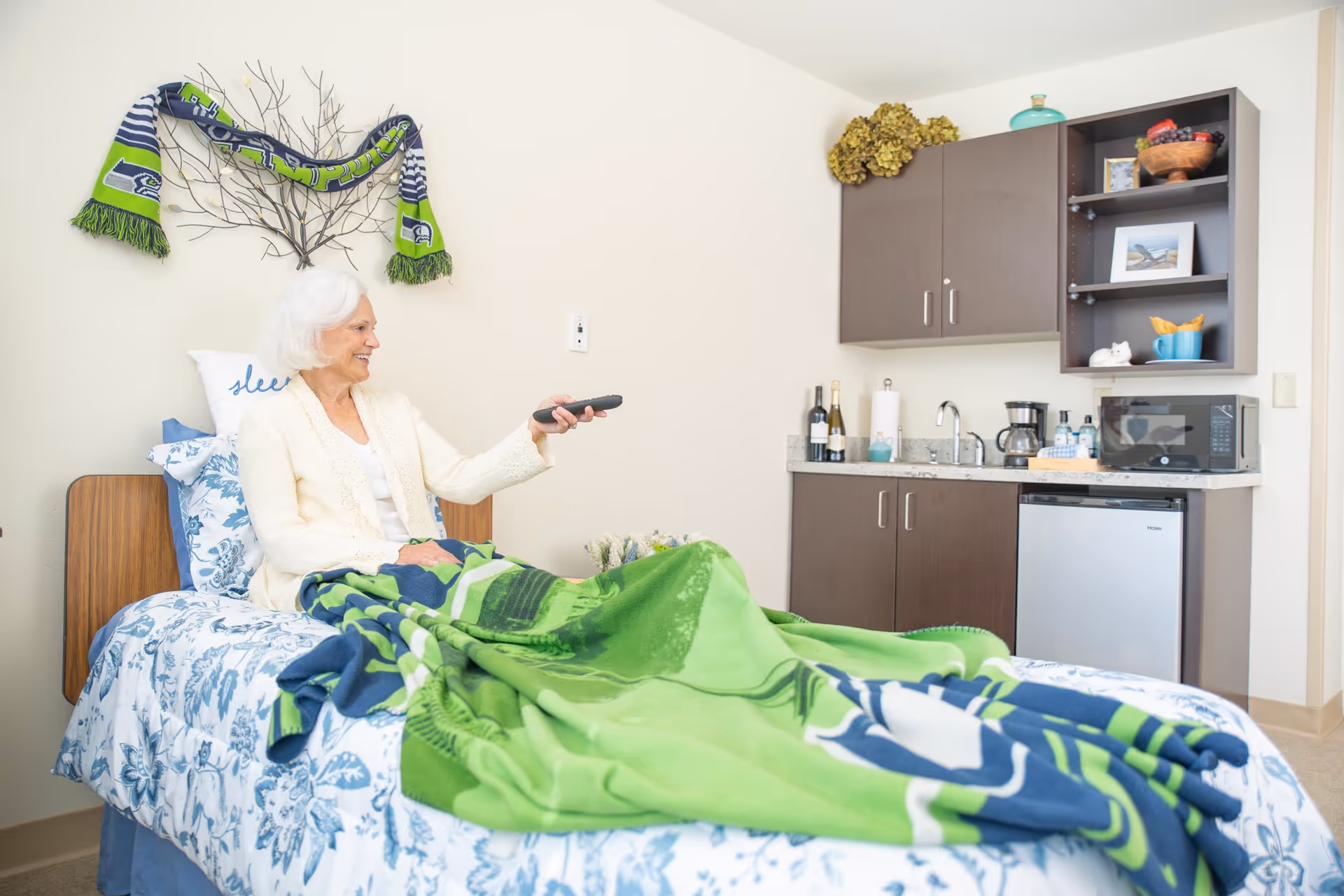 An elderly woman sits on a bed holding a TV remote in a bright assisted-living room with a small kitchenette and cabinets behind her.