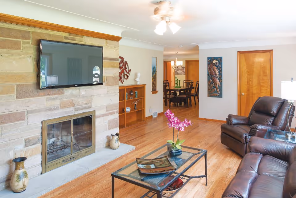 A bright living room with hardwood floors featuring a stone fireplace with a mounted flat-screen TV above it. There are two dark brown leather recliners and a glass coffee table with a pink orchid and decorative items. In the background, a dining area with a wooden table and chairs is visible, along with wall art and a ceiling fan with lights.