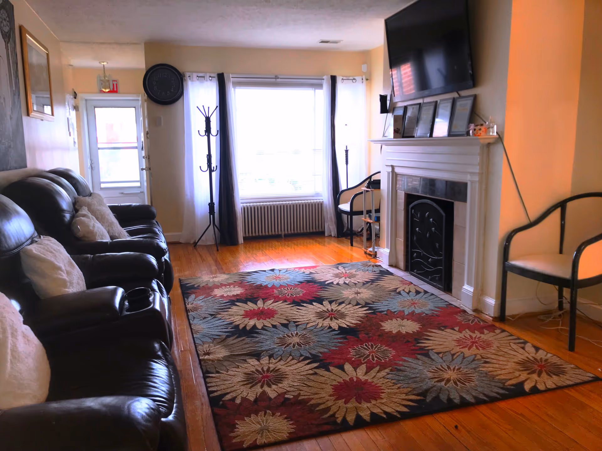 A cozy living room with three dark leather recliners on the left side, each with a light-colored pillow. A colorful floral area rug covers the wooden floor. On the right side, there is a white fireplace with a black decorative screen and several framed certificates or pictures on the mantel. Above the fireplace is a mounted flat-screen TV. A window with white and dark curtains is at the far end of the room, next to a coat rack and a chair. The walls are painted a light beige color.