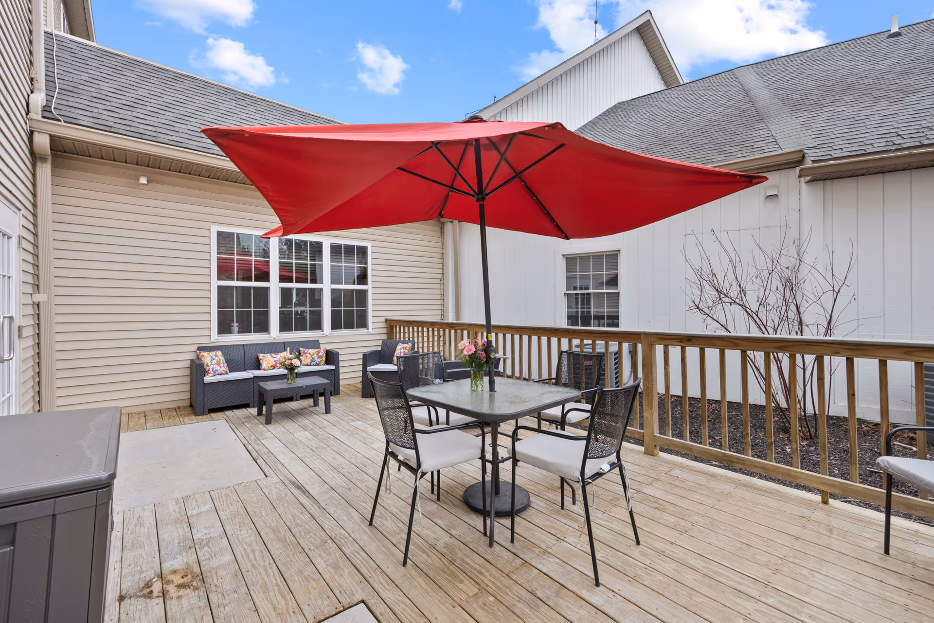 Outdoor wooden deck area with a red patio umbrella over a glass table surrounded by four chairs. There is a small seating area with a black couch and two chairs with colorful cushions against the beige siding of the building. A wooden railing encloses the deck, and the sky is partly cloudy.