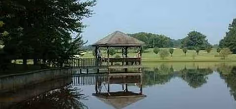 Wooden gazebo beside a tranquil pond with a reflection and a grassy park.