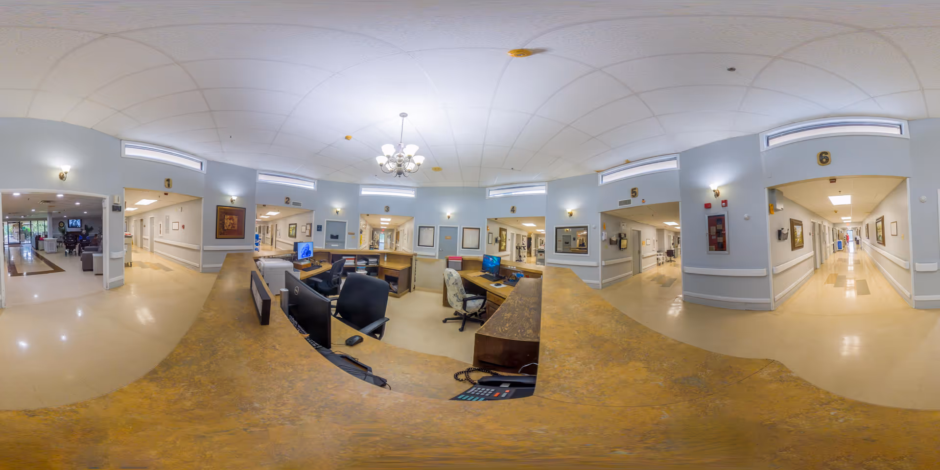 Wide panoramic view of a nursing and rehab center's interior nurse station area with numbered rooms and hallways extending on both sides. The nurse station has multiple computer monitors and office chairs. The walls are painted light blue with white trim, and there are framed pictures and wall sconces. The floor is a light beige color, and a chandelier hangs from the ceiling in the center.