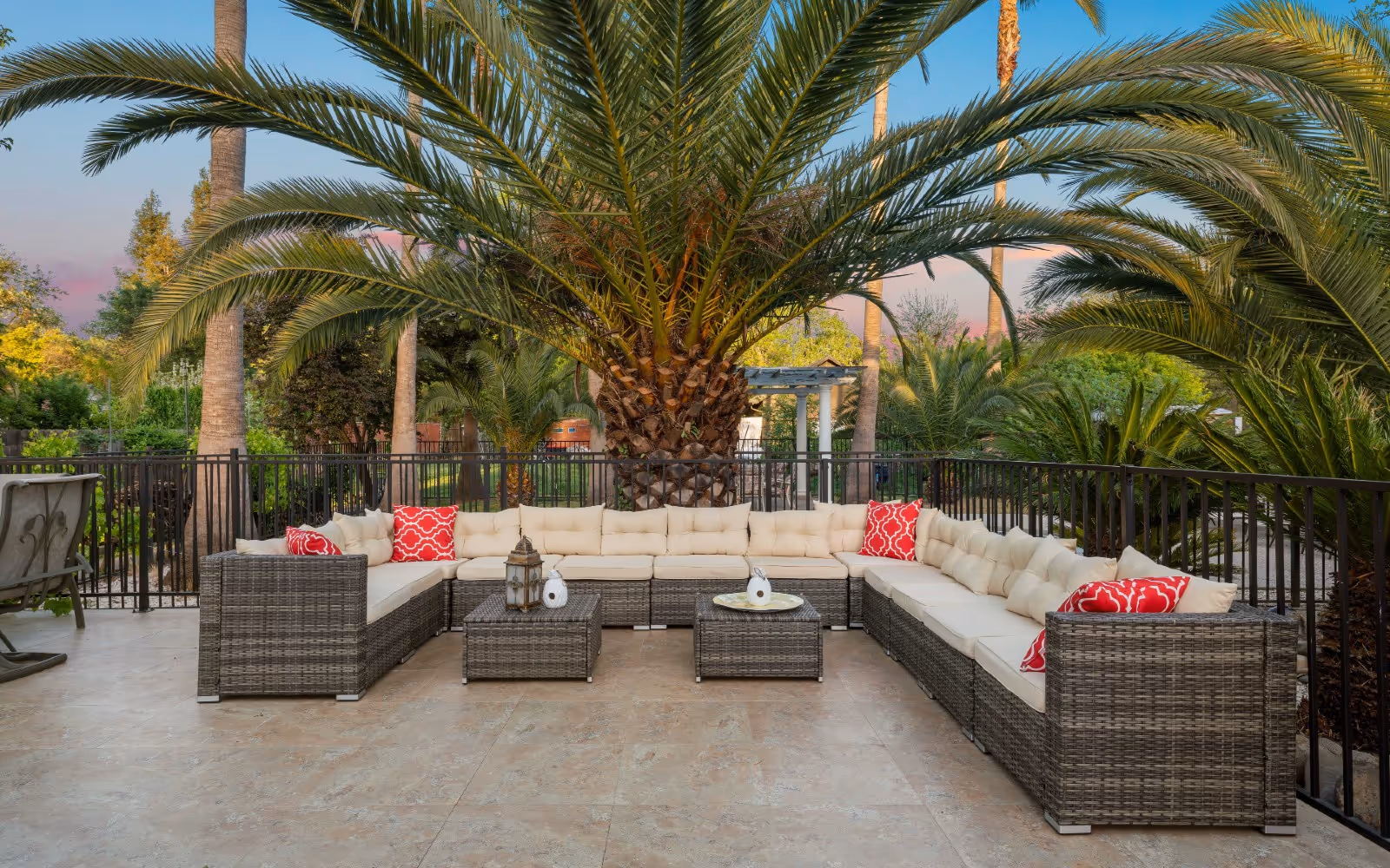 Outdoor patio area with a large U-shaped wicker sectional sofa with beige cushions and red patterned throw pillows, two matching wicker coffee tables with decorative items, surrounded by palm trees and greenery under a clear sky.