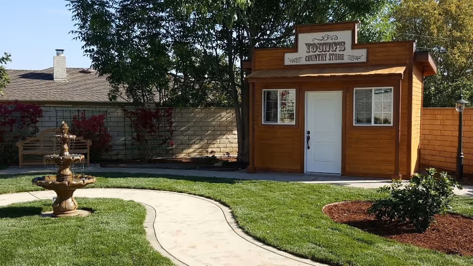 A small wooden building labeled 'Young's Country Store' with a white door and two windows, situated in a garden area with a curved concrete pathway, green grass, a multi-tiered water fountain, a bench, and trees in the background.