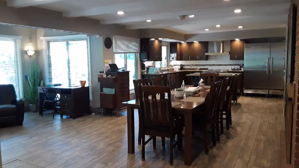 A spacious interior room featuring a large wooden dining table with multiple chairs, a modern kitchen area with stainless steel refrigerator, dark wood cabinets, and a countertop. The room has wooden flooring, large windows with blinds allowing natural light, and a small desk with a chair near the windows.