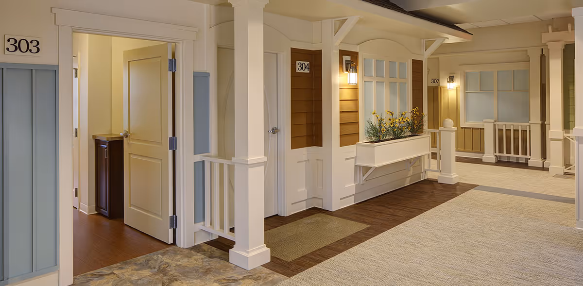 Indoor hallway of a senior living facility designed to resemble a residential street with apartment doors numbered 303, 304, and 307. The area features warm lighting, a flower box with yellow flowers under a window, and a mix of carpet and wood flooring.