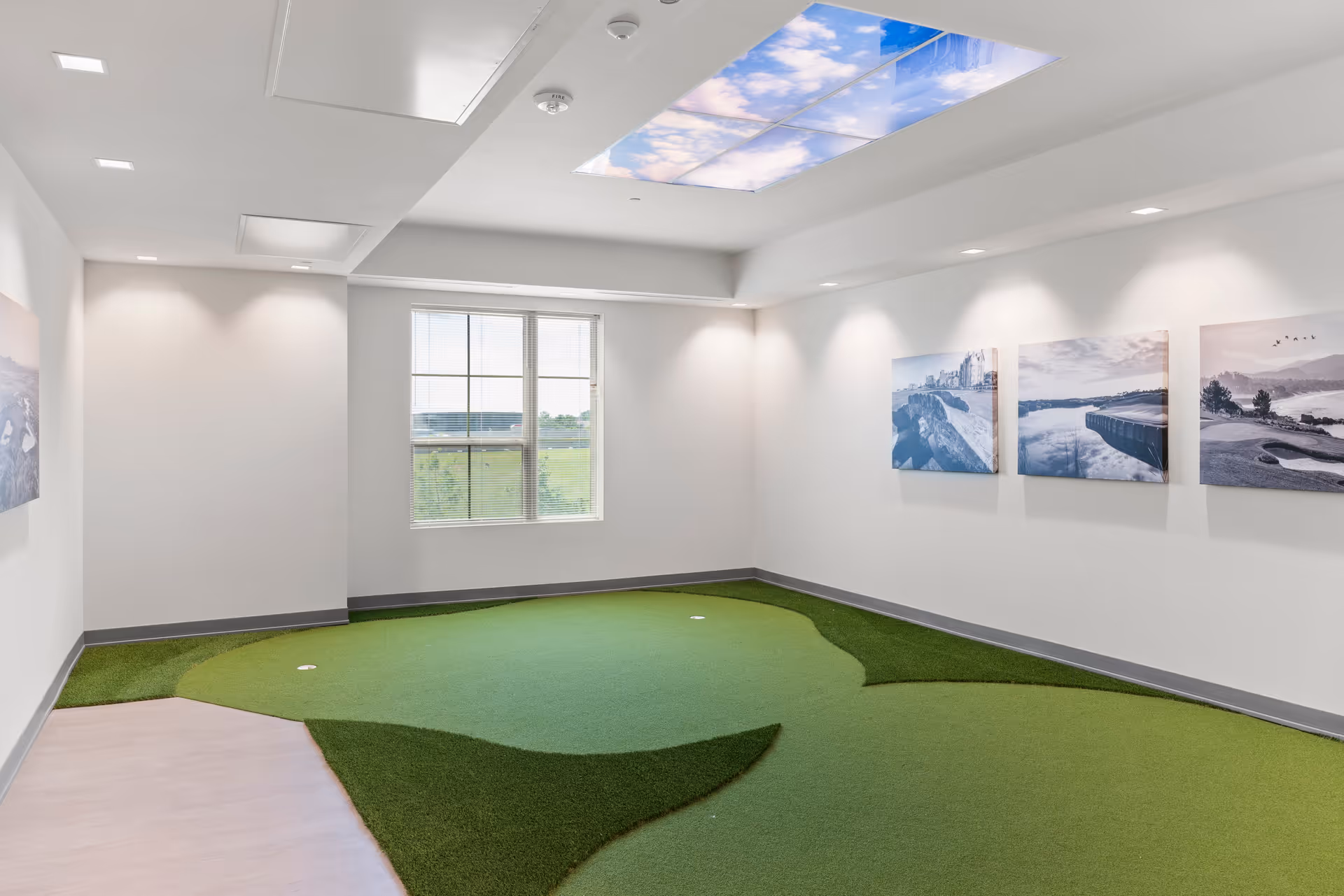 Indoor room with artificial putting green on the floor, white walls with three landscape pictures, a window showing outdoor greenery, and a ceiling panel displaying a blue sky with clouds.