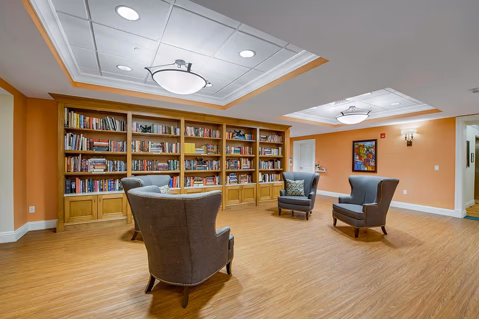 A cozy reading room with wooden flooring, orange walls, and a large wooden bookshelf filled with books. Four gray upholstered armchairs with decorative pillows are arranged in a semi-circle in the center of the room. The ceiling has recessed lighting and two large modern light fixtures. A colorful framed painting hangs on the wall near a doorway.