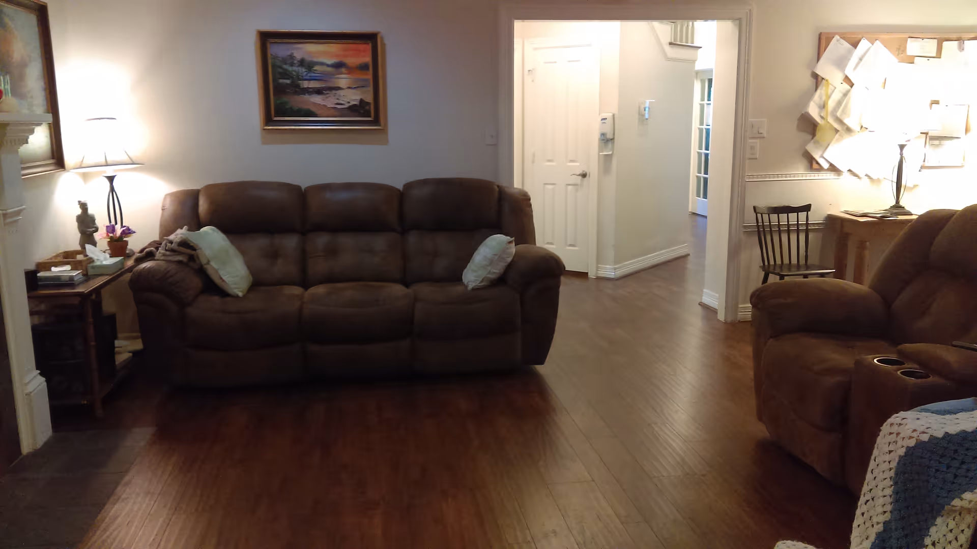 A cozy living room with a brown three-seater sofa and a matching reclining chair. There are two pillows on the sofa and a side table with a lamp, books, and a small statue. The room has wooden flooring and light-colored walls with framed artwork. A hallway with a white door and a bulletin board with papers is visible in the background.