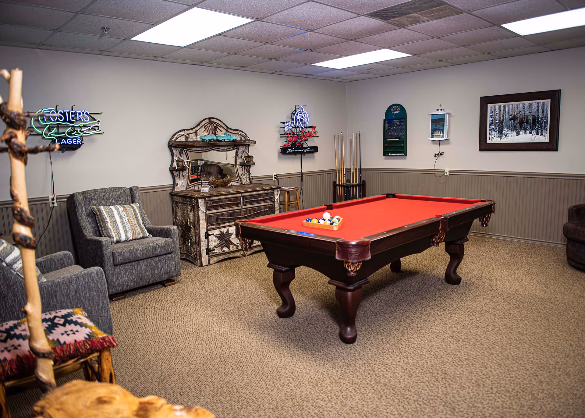 A recreational room with a red felt pool table in the center, two gray armchairs with a striped pillow, a rustic wooden cabinet with a mirror, pool cues in a rack, neon signs on the walls, and a framed picture of a forest scene.