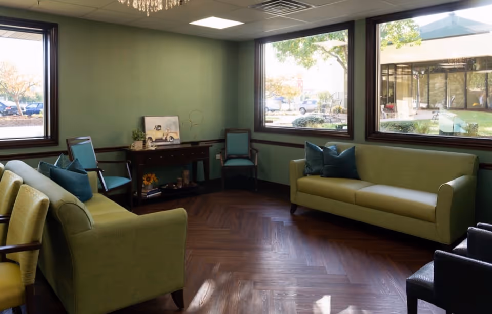 A cozy living room with green walls and large windows letting in natural light. The room features two green sofas with blue pillows, several chairs, a wooden console table with decorative items, and dark wood herringbone flooring. Outside the windows, trees and parked cars are visible.