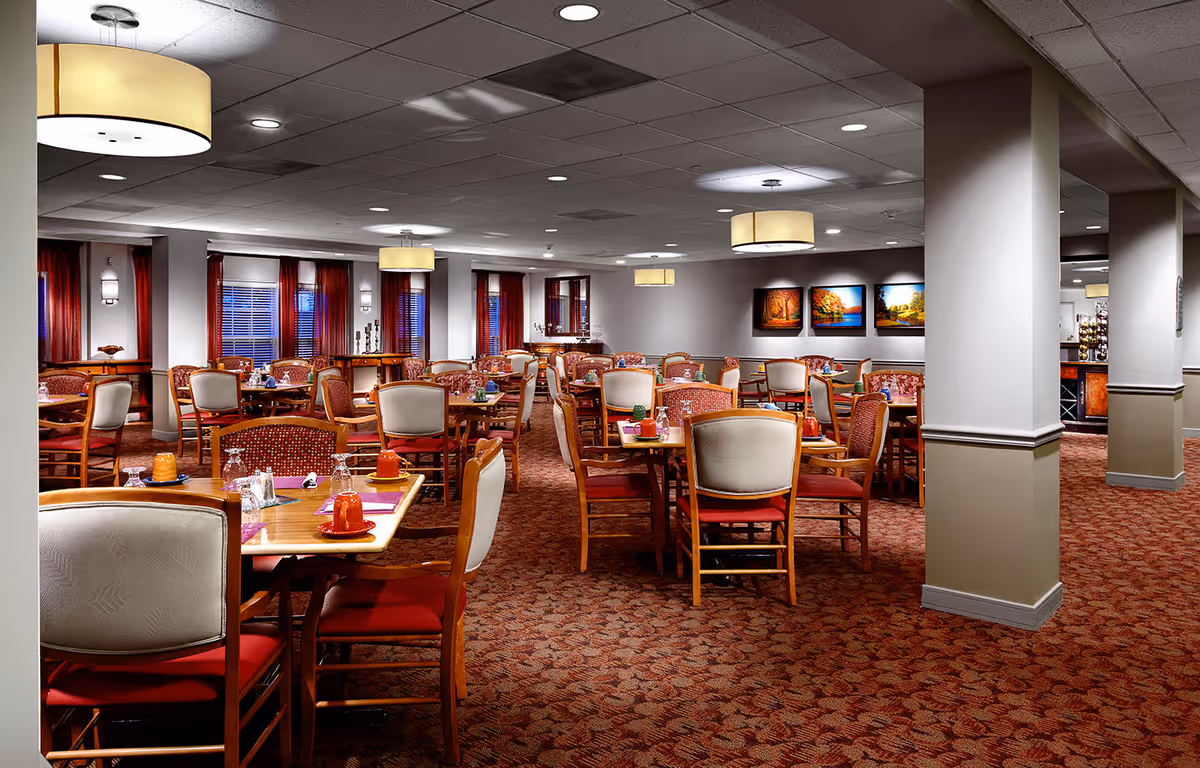 A spacious dining room with multiple wooden tables and chairs arranged neatly. The tables are set with placemats, cups, and glasses. The room has carpeted flooring with a patterned design, white walls with red curtains on the windows, and several ceiling lights providing warm illumination. There are framed landscape paintings on one wall and a buffet area visible in the background.