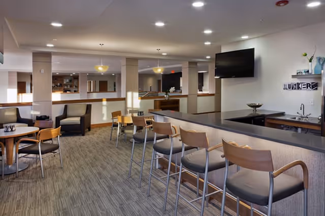 Spacious communal dining and lounge area with a long bar counter and high stools, small dining tables and chairs, lounge seating, and a wall-mounted TV.