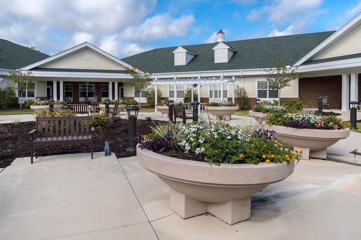 Outdoor courtyard area of Rice Estate featuring large round concrete planters filled with colorful flowers, several wooden benches, small trees, and a pergola in front of a single-story building with a green roof and beige siding under a partly cloudy sky.